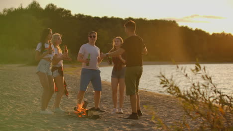 compañía de cinco jóvenes están bailando en pantalones cortos y camisetas alrededor de la hoguera en la fiesta al aire libre con cerveza. están sonando y bebiendo cerveza y disfrutando de la noche de verano al atardecer cerca del bosque.