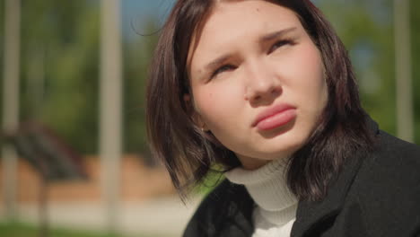 close-up of woman outdoors squinting in bright sunlight, wearing a black coat and white sweater, holding a phone, thoughtful expression with blurred background of greenery and trees