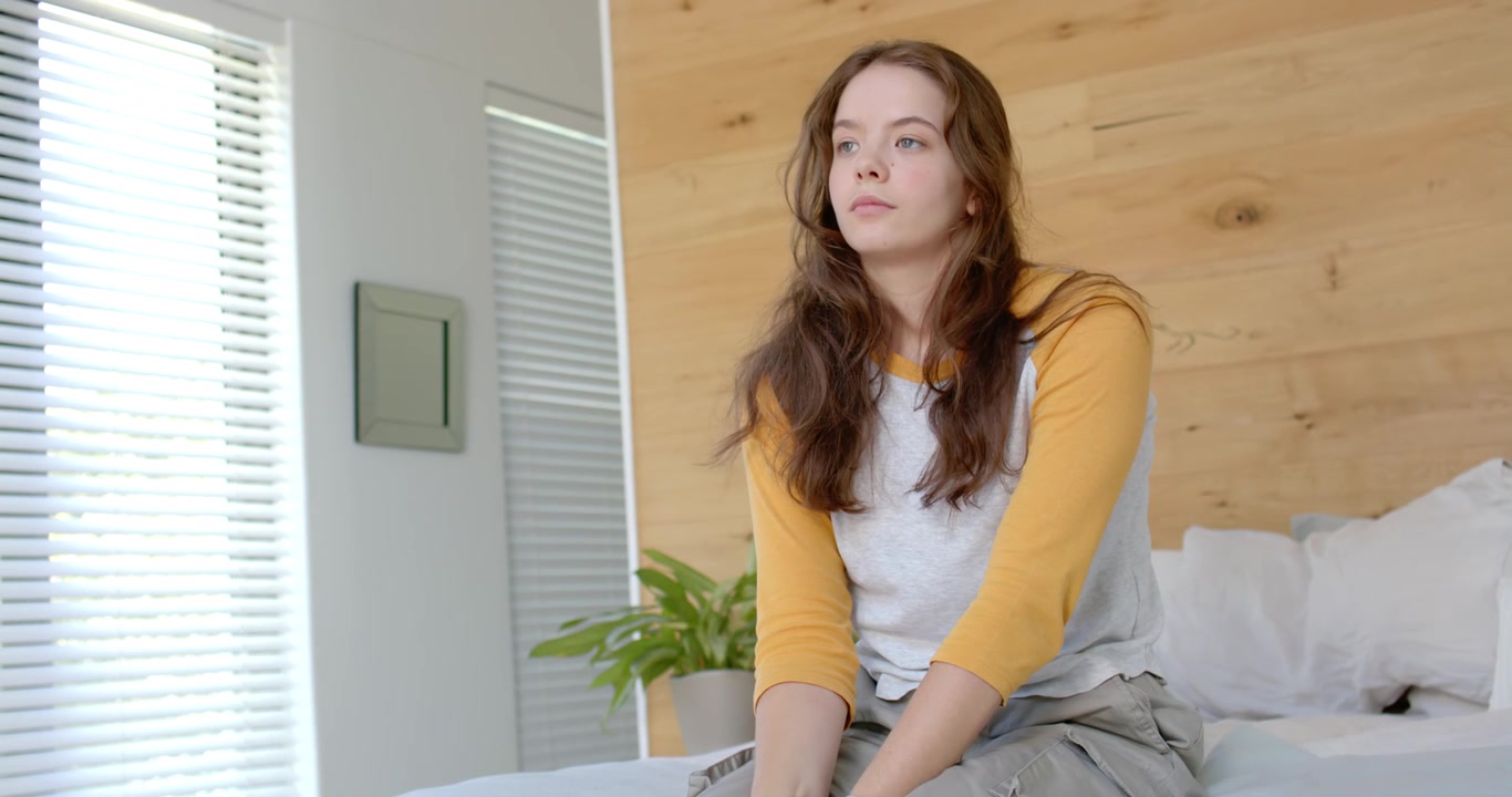 Mulher sentada na beira da cama, virando a cabeça e olhando para a direita, contemplando, vestindo uma blusa raglan mostarda