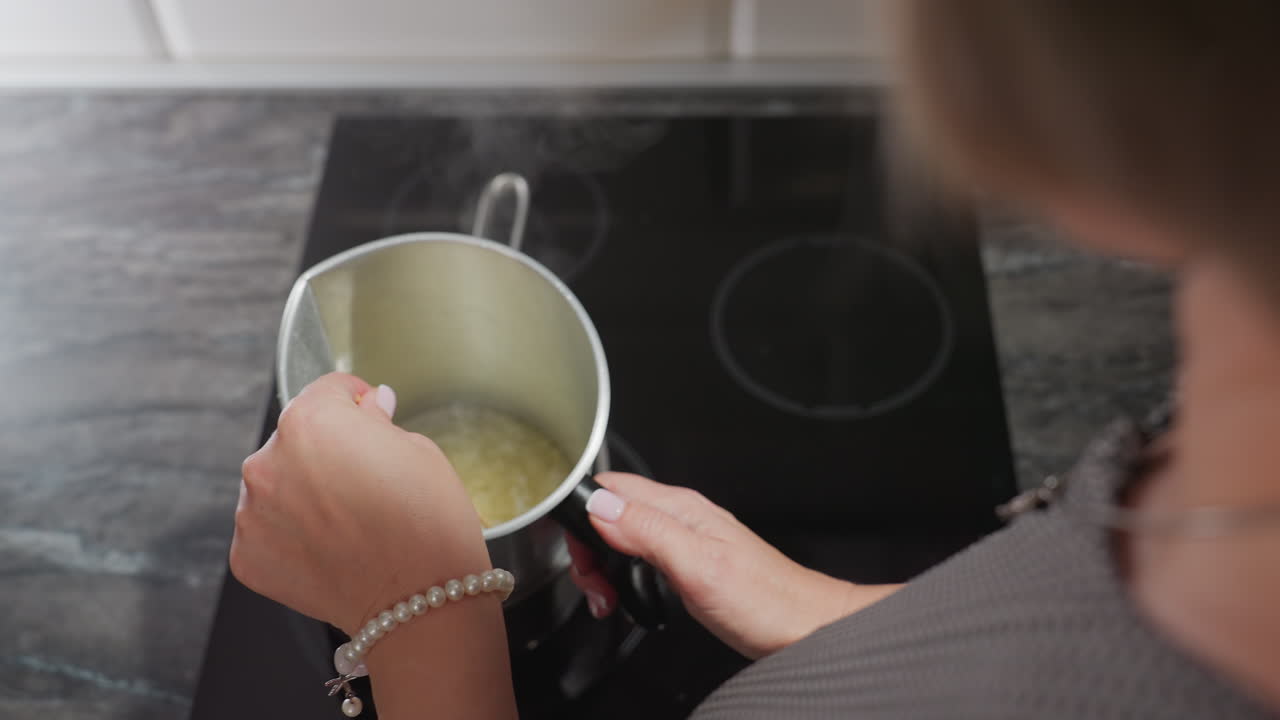 Fair skinned creative woman stirs sugar mixture with left hand over heated burner as steam rises from metal container, capturing precision, and creative kitchen process with elegance