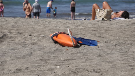 Orangefarbener-Rettungsring-Und-Blaue-Flossen-Liegen-Auf-Dem-Sandstrand,-Symbole-Für-Sicherheit-Im-Wasser-Und-Abenteuer-Unter-Entspannten-Strandbesuchern-Unter-Der-Sommersonne-Mit-Einem-Mann,-Der-In-Sandalen-Läuft