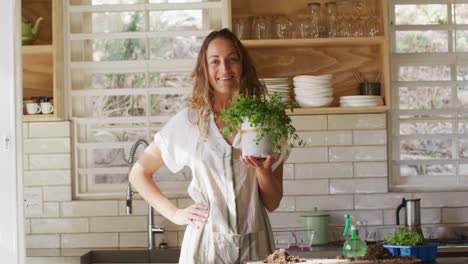 Retrato-De-Una-Mujer-Caucásica-Sonriente-Sosteniendo-Una-Planta-En-Maceta,-Parada-En-La-Soleada-Cocina-De-La-Cabaña