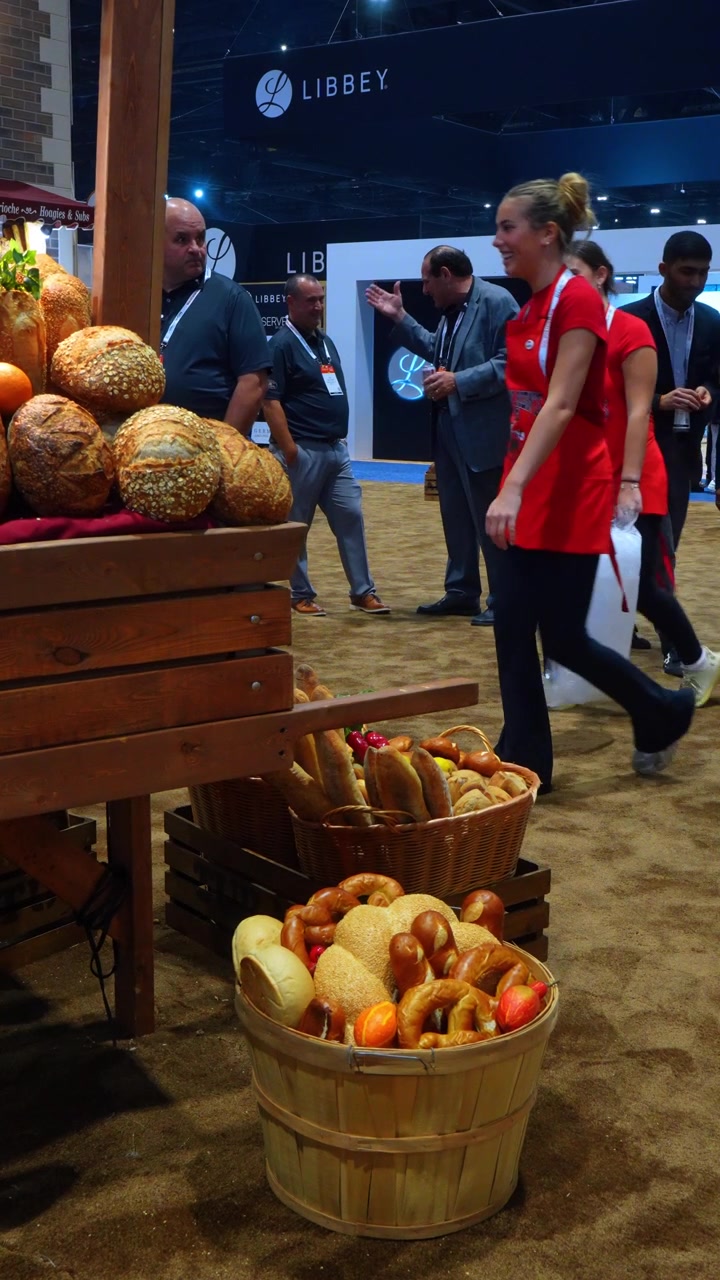Chicago, USA, 9 maggio 2026: Esposizione di pane accanto a visitatori d'affari a una fiera di Chicago. La vista verticale dell'esposizione mostra pagnotte in cesti, carrello di legno, partecipanti e segnaletica dello stand con marchio
