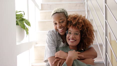 Portrait-of-happy-diverse-lesbian-female-couple-on-stairs-and-embracing-at-home-in-slow-motion