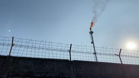 Industrial-gas-plant-tower-emitting-Co2-in-atmosphere,-low-angle-behind-fence