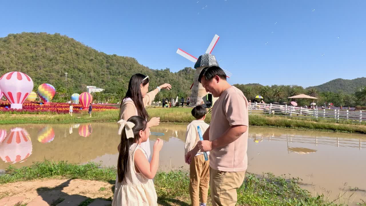 ครอบครัวชาวเอเชียที่มีความสุขเป่าฟองสบู่อยู่ริมสระน้ำ โดยมีกังหันลมและฉากหลังเป็นภูเขา