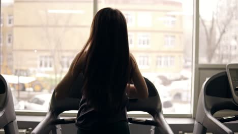 backside footage in slow motion of a sportive girl with long brunette hair running on a treadmill. indoors.