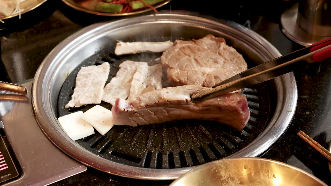 Hand uses tongs to flip pork ribs on tabletop Korean barbecue grill under warm lighting