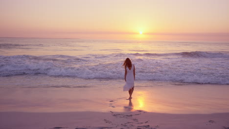 beautiful woman wearing white dress walking along shore line on  beach at sunset in slow motion red dragon