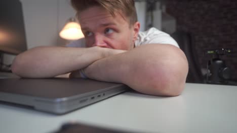 a young man with dark blonde hair leans over his desk and computer completely exhausted before he gets up and walks away