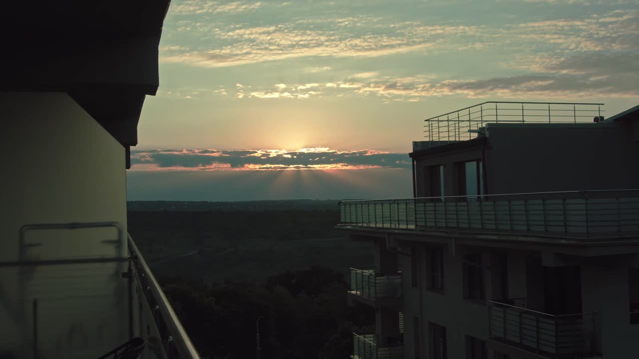 impresionante vista de la puesta de sol desde un balcón moderno, con el cielo sereno de la noche, los rayos del sol rompiendo a través de las nubes, y el paisaje urbano pacífico