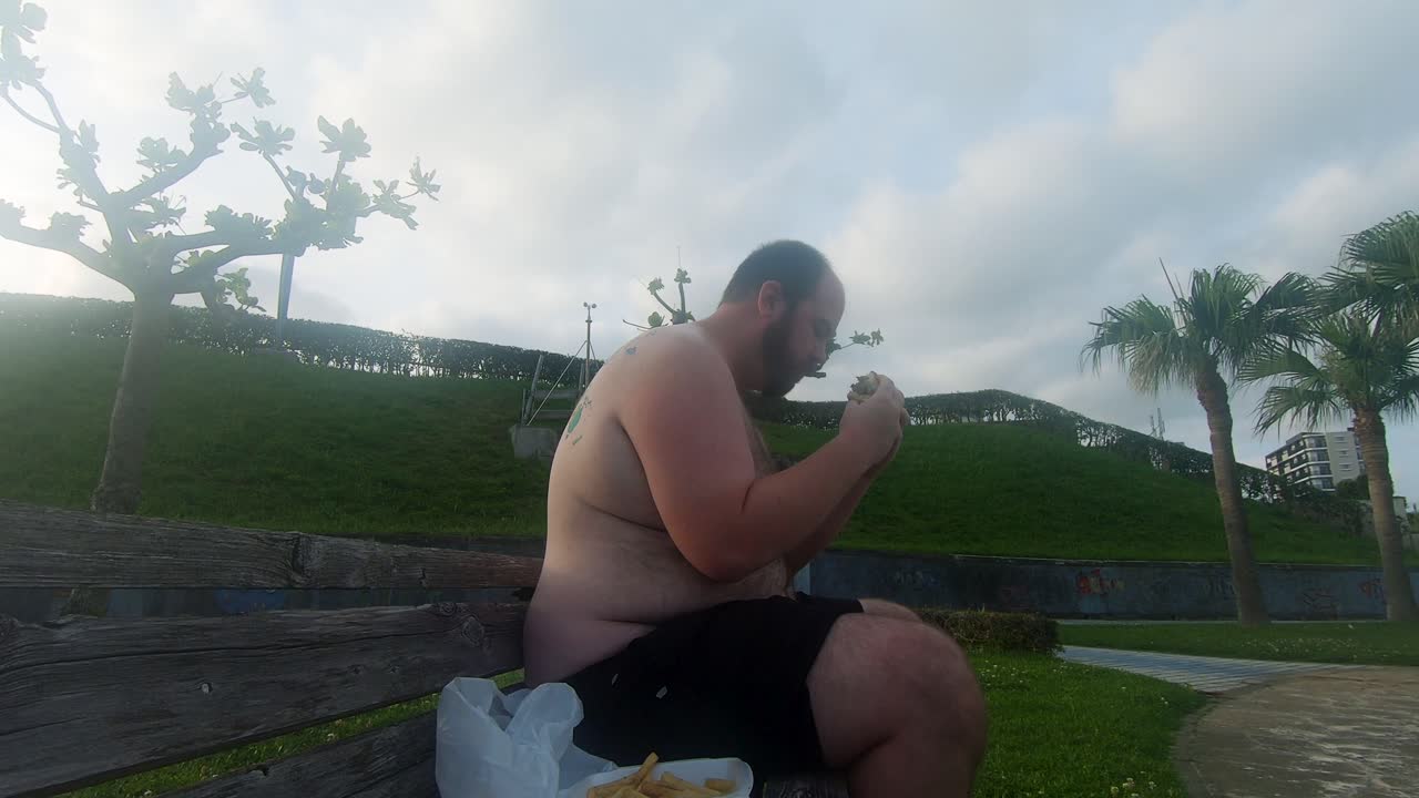 el hombre obeso que come una hamburguesa grasienta en un banco del parque se inclina hacia atrás y luego se frota el estómago gordo y peludo