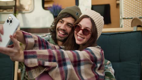 A-happy-brunette-girl-in-round-glasses-and-a-plaid-shirt-together-with-her-brunette-boyfriend-in-a-green-hat-with-stubble-takes-a-selfie-against-the-backdrop-of-a-trailer-and-a-green-chair-during-their-picnic-outside-the-city