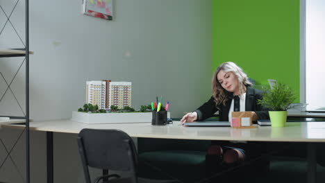 real estate agent sits at modern desk on phone call with client preparing for workday by opening laptop, architectural model around, green plant, file stacks, and colorful stationery in bright office