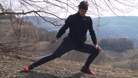 a dynamic outdoor shot of an individual in a black long‑sleeve and striped black pants, performing a lunge stretch with red shoes amid autumn hills and bare trees.