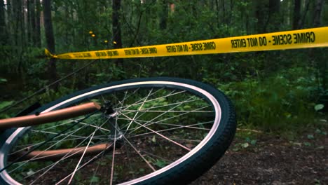 low angle eerie and dark footage of a crashed bicycle in a forest with its wheel spinning slowly and yellow crime scene do not cross tape in the background moving in the wind on a forest muddy road.