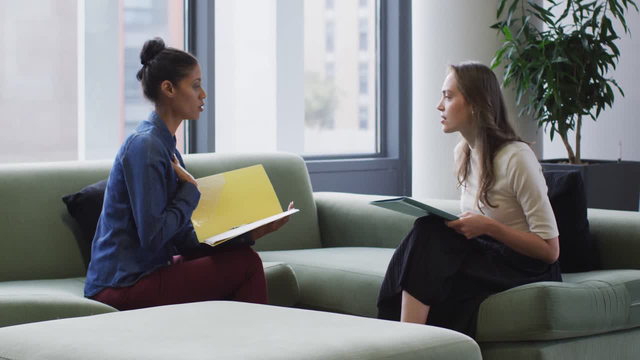 To mangfoldige kvindelige arbejdskolleger sidder på en sofa, holder dokumenter og taler.