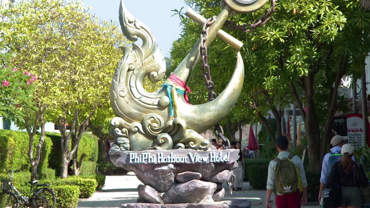 Du khách đi bộ qua khu vườn nghỉ dưỡng ngập nắng, ngang qua một bức tượng mỏ neo vàng lớn ở Thái Lan.