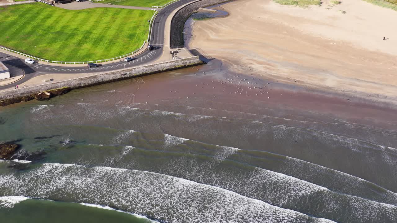 Drone glides above West Sands Beach, capturing waves, shoreline, green park, and waterfront road