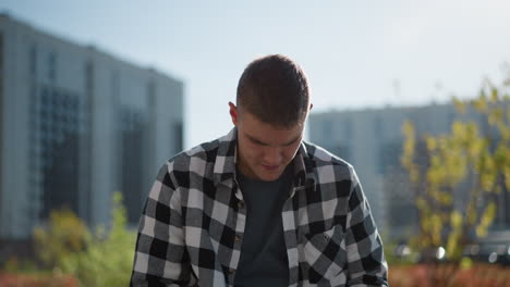 young artist in checkered shirt looks down while seated outside taking medication with blurred background of city buildings, parked cars, green trees and signpost under bright daylight atmosphere
