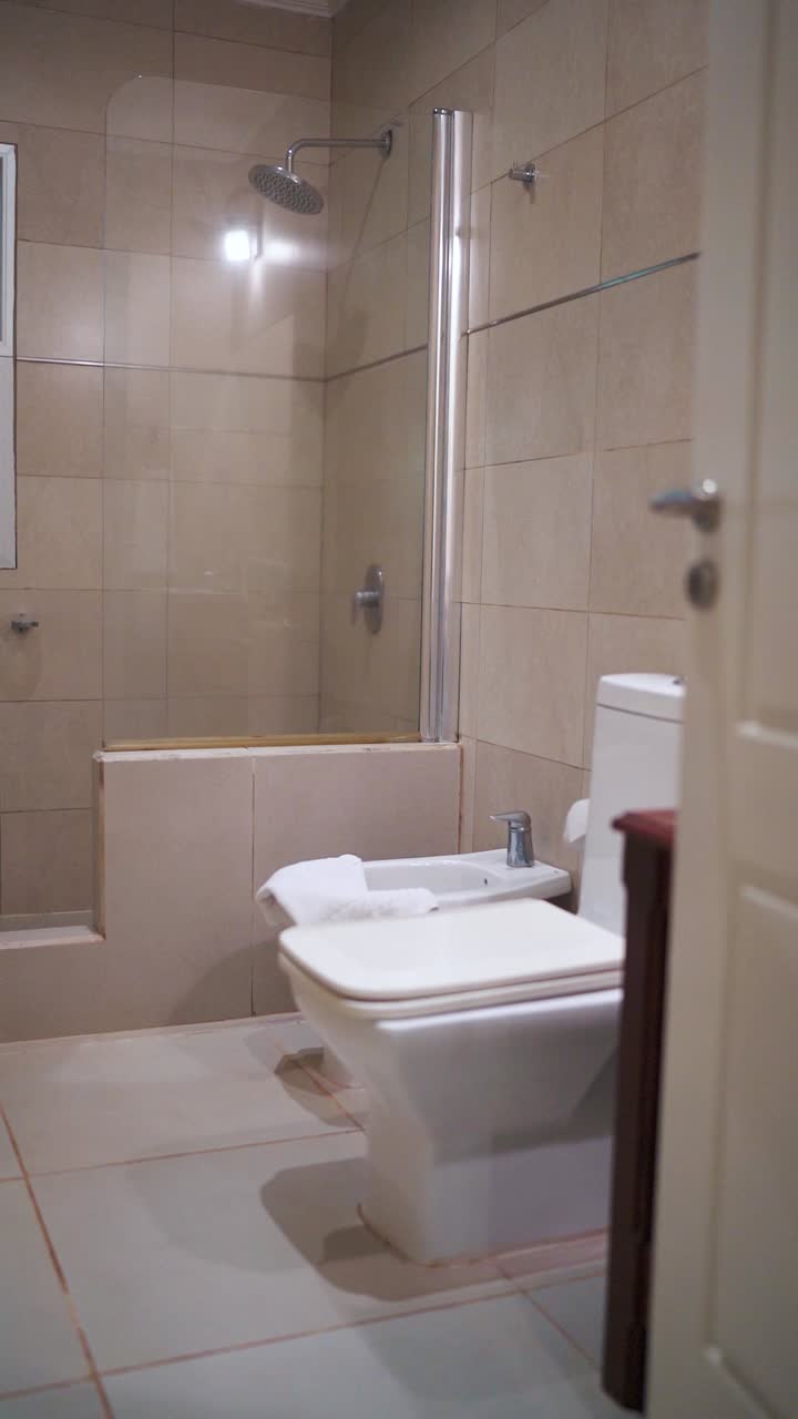 Slow right moving vertical camera shot of rest room at hotel, with light beige color tiles and white ceramic toilet and bidet.