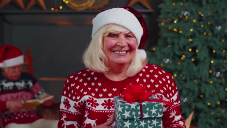 Abuela-Mayor-Con-Suéter-Festivo-Presentando-Caja-De-Regalo-De-Navidad,-Sonriendo,-Mirando-A-La-Cámara