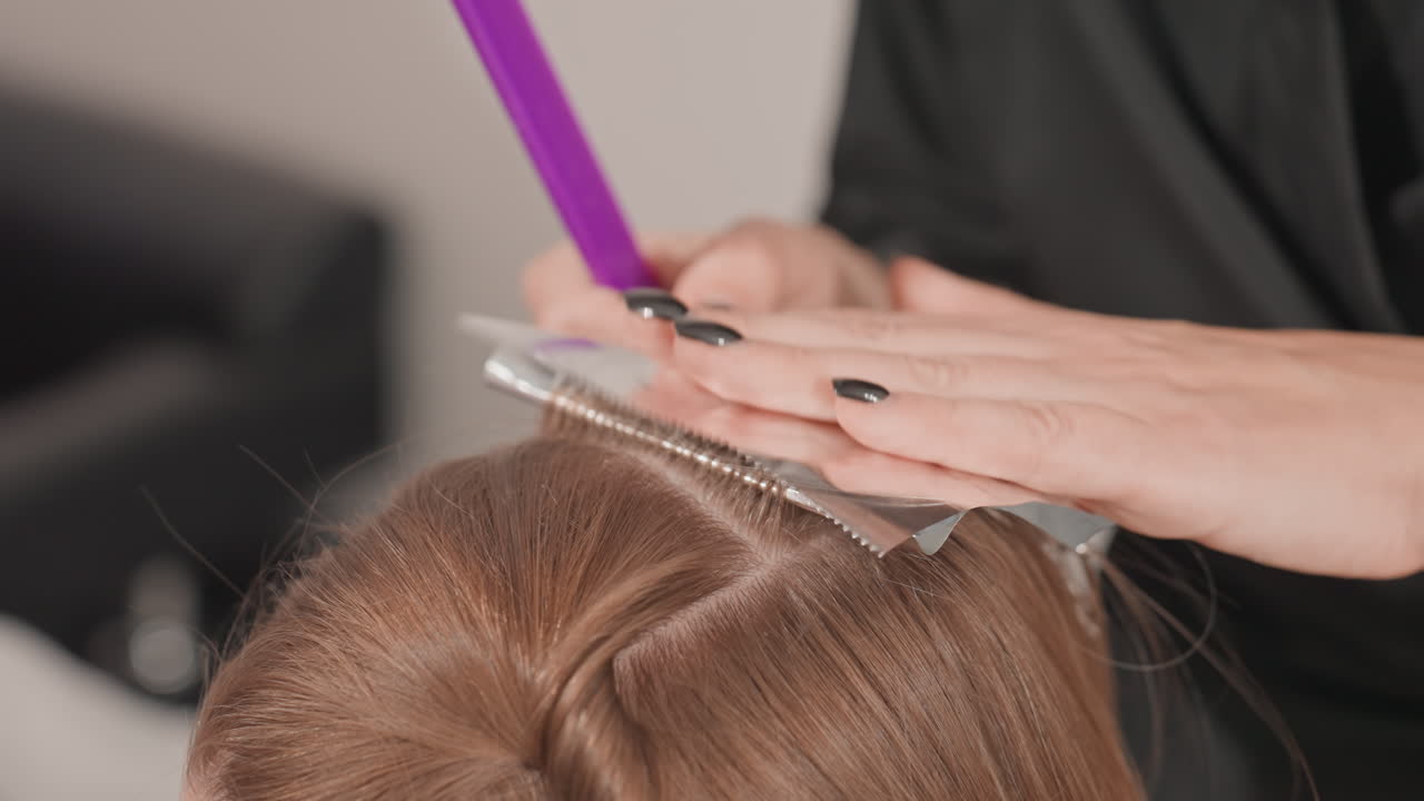 Parrucchiere che fa colpi di sole applicando il colore ai capelli castani, mani in primo piano che posizionano un foglio di alluminio e pettinano una sezione, manicure nera e pettine a coda viola visibili, colorista professionista all'opera