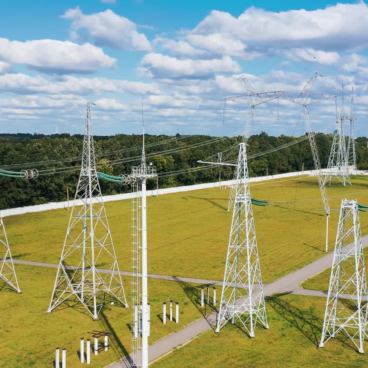High voltage steel pylons on green field. Power transmission lines at sunlight. White electricity towers in summer day