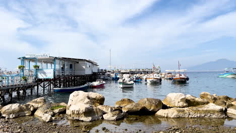 A-waterfront-scene-with-moored-boats-beside-a-pier-with-buildings,-under-a-clear-sky,-and-a-mountain-backdrop