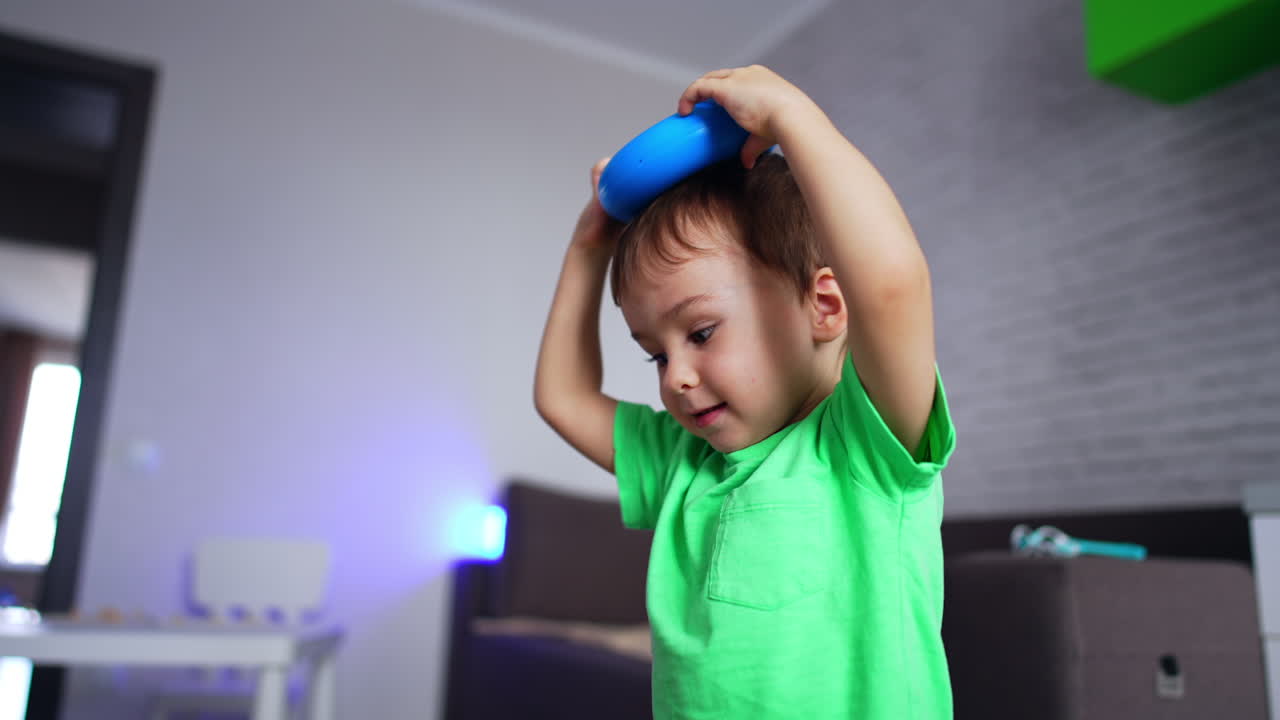 Cute toddler in light blue t-shirt puts the ring from a toy pyramid on his head. Happy kid having fun at home. Blurred backdrop.