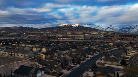 Sunset-time-lapse-over-a-suburb-and-business-community-below-the-snowy-mountains---aerial-hyper-lapse