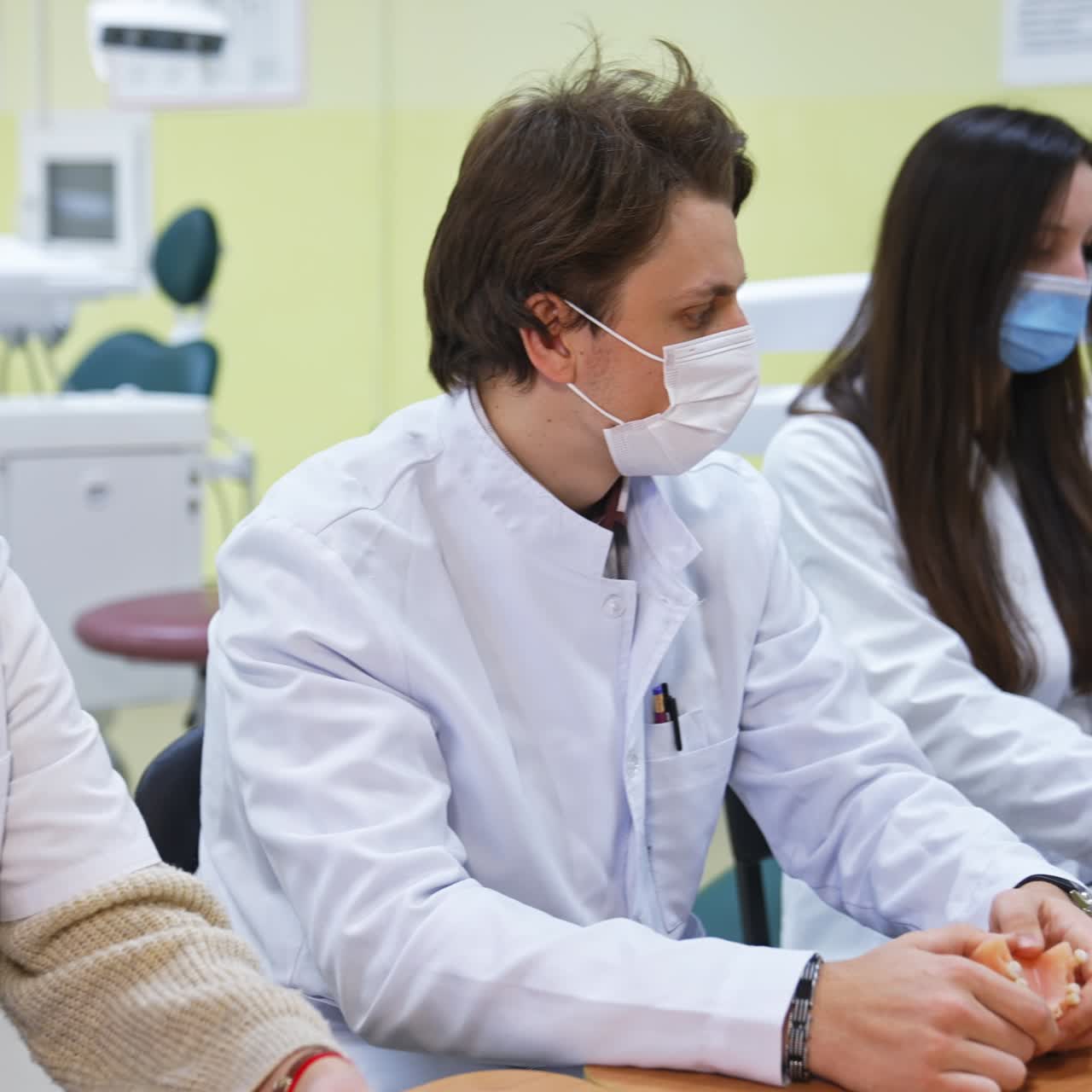 Students having a lecture sitting at the desk in classroom. Man standing in front of them and showing rubber dam. Auditory in blur at the backdrop