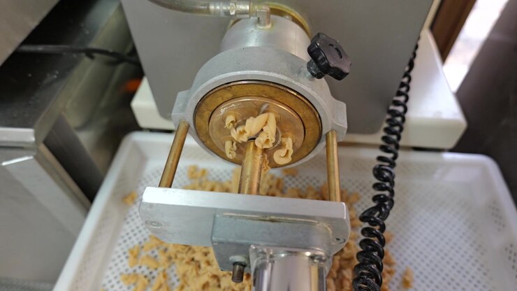 Primo piano dall'alto di una macchina per l'estrusione della pasta che forma pezzi di pasta fresca sopra un vassoio perforato in una cucina di produzione.