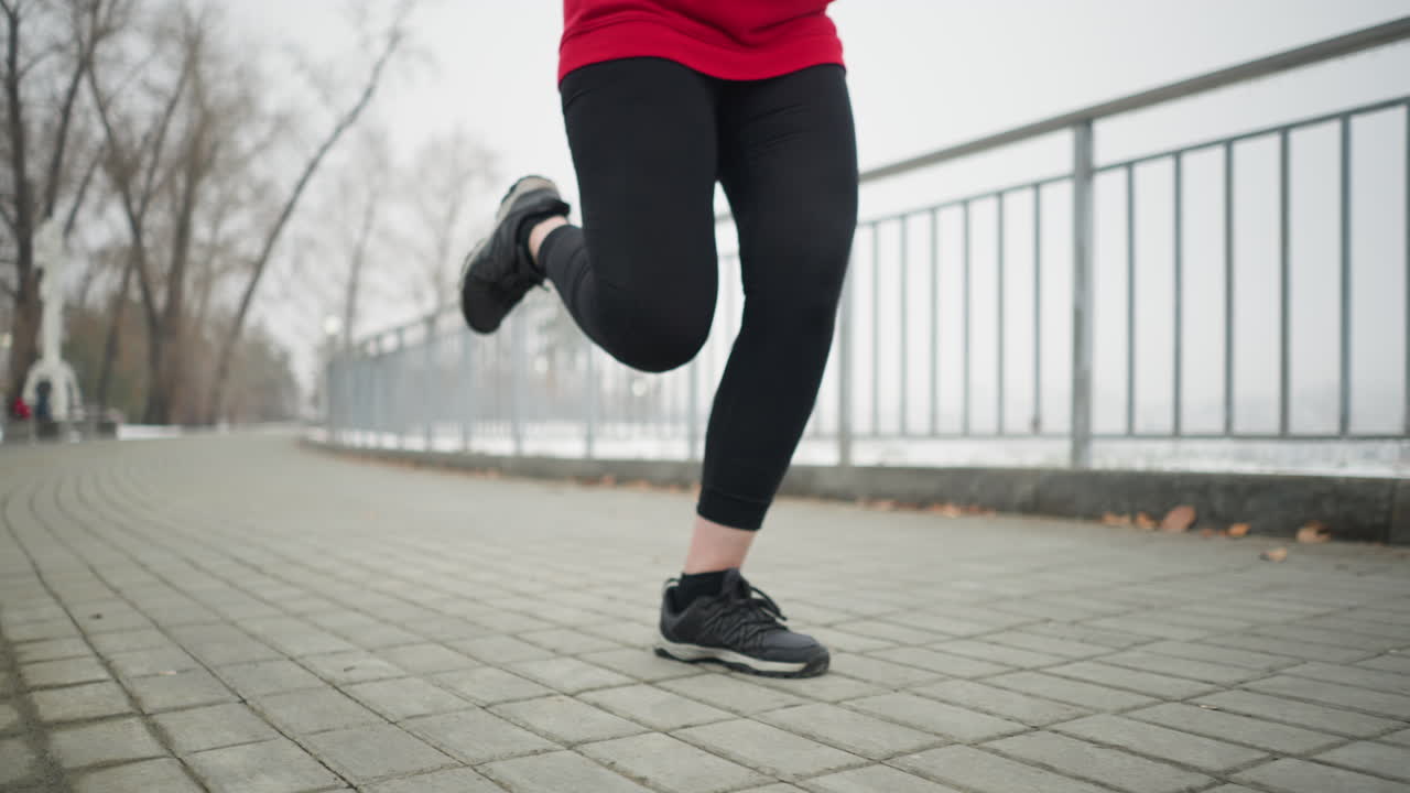 vista de ángulo inferior de atleta en zapatillas de deporte negras y leggings corriendo a lo largo de la barandilla de hierro del puente rodeado por la atmósfera de invierno brumosa, vista parcial del paisaje urbano, fondo bordeado de árboles