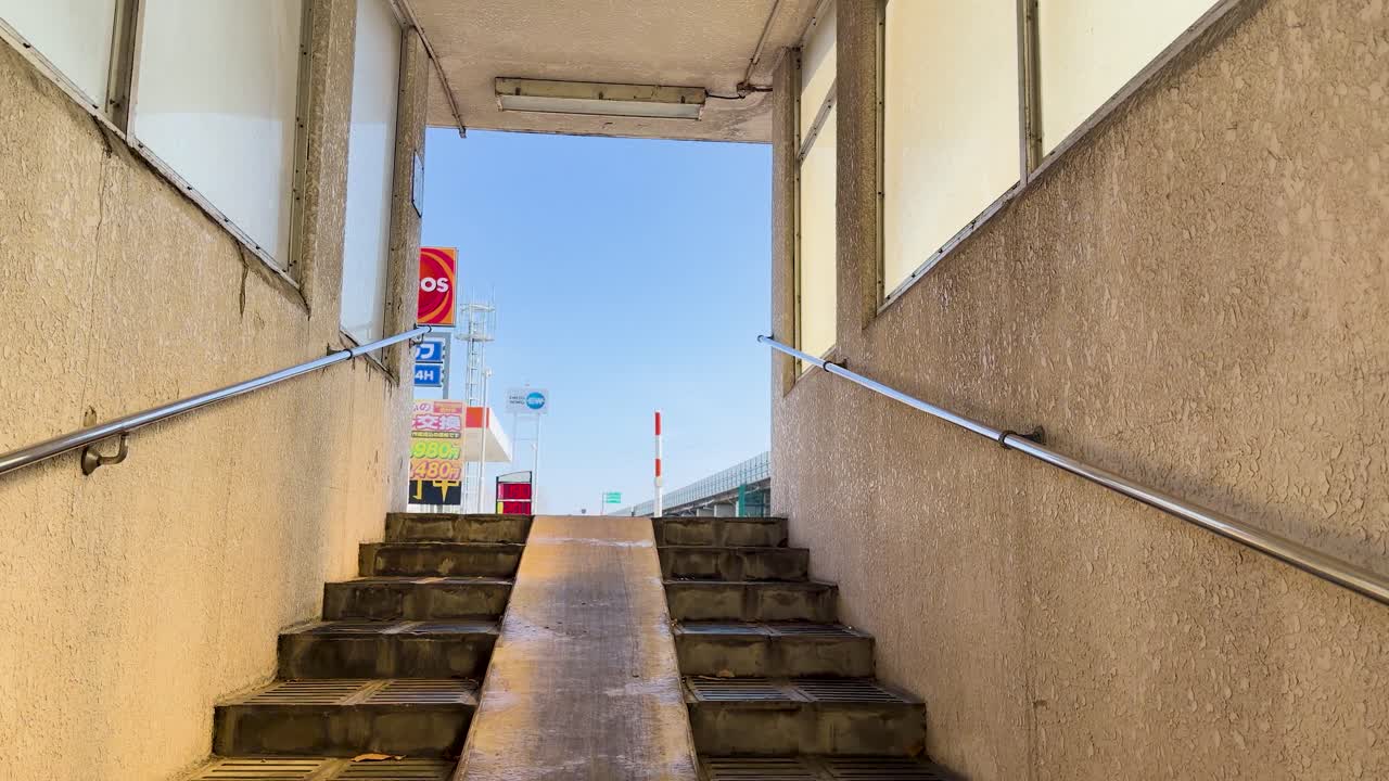 Inquadratura fissa dal basso di scale di cemento e rampa metallica che conducono verso una luminosa strada cittadina.