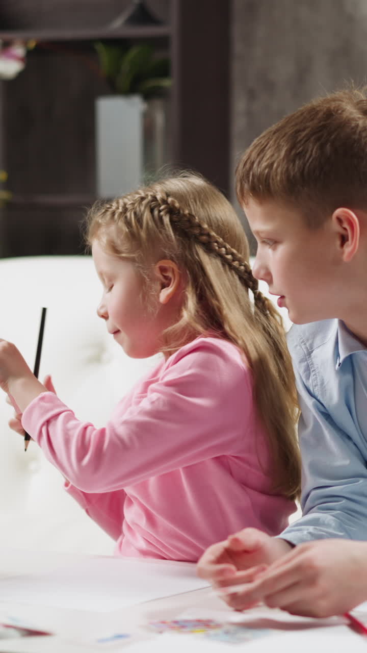 Blonde junior schoolgirl takes card with letter and hides from brother doing English language homework together at small table in living room slow motion