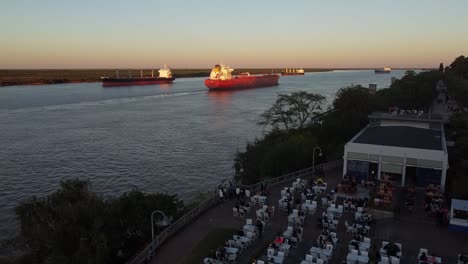 Vista-Aérea-Del-Restaurante-En-La-Costa-Del-Río-Con-El-Paraná-Al-Fondo-Y-Varios-Barcos-Navegando,-En-Una-Tarde-De-Verano