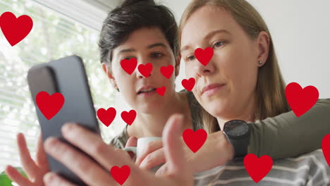 animación de corazones sobre una pareja de mujeres caucásicas usando un teléfono inteligente.