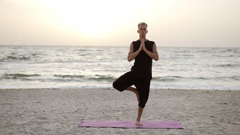 portrait of a young man who practices yoga on a sports mat, standing on one leg at dawn. performing a specific exercise