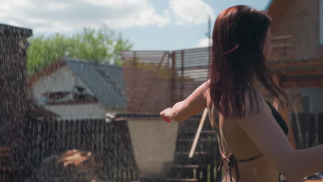 excited woman with back tattoo jumps under refreshing water spray from hose in backyard droplets glistening in sunlight splashing and laughing amid green lawn by wooden fence