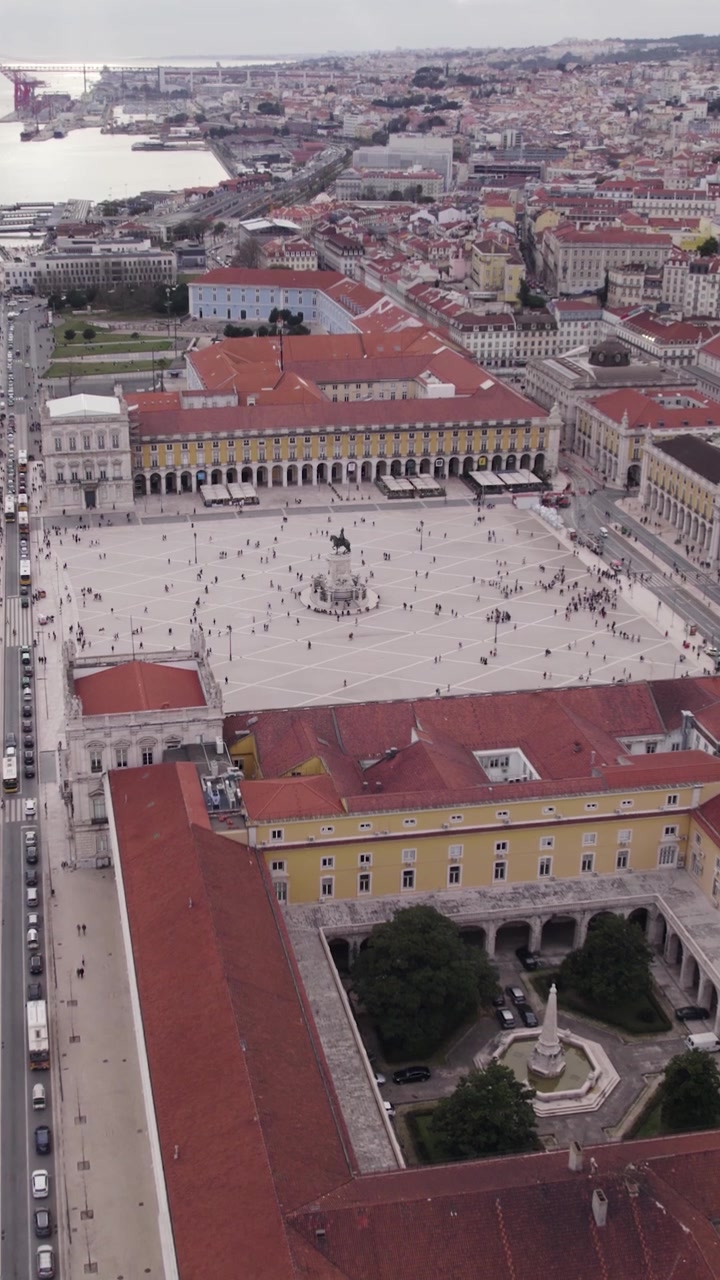 Ripresa verticale con drone che avanza sopra il Terreiro do Paço, mostrando la piazza storica, la statua, l'architettura circostante e lo skyline di Lisbona