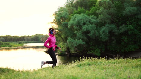 a girl in a pink jacket and black pants runs near the river in headphones preparing for the marathon