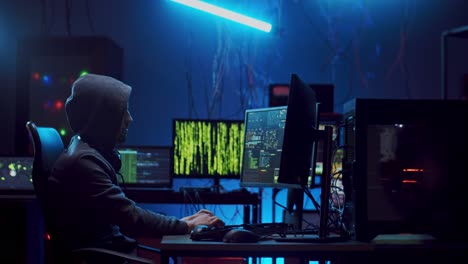 side view on the anonymous male cyber criminal in hood hacking data and coding viruses at the computer in the dark at night