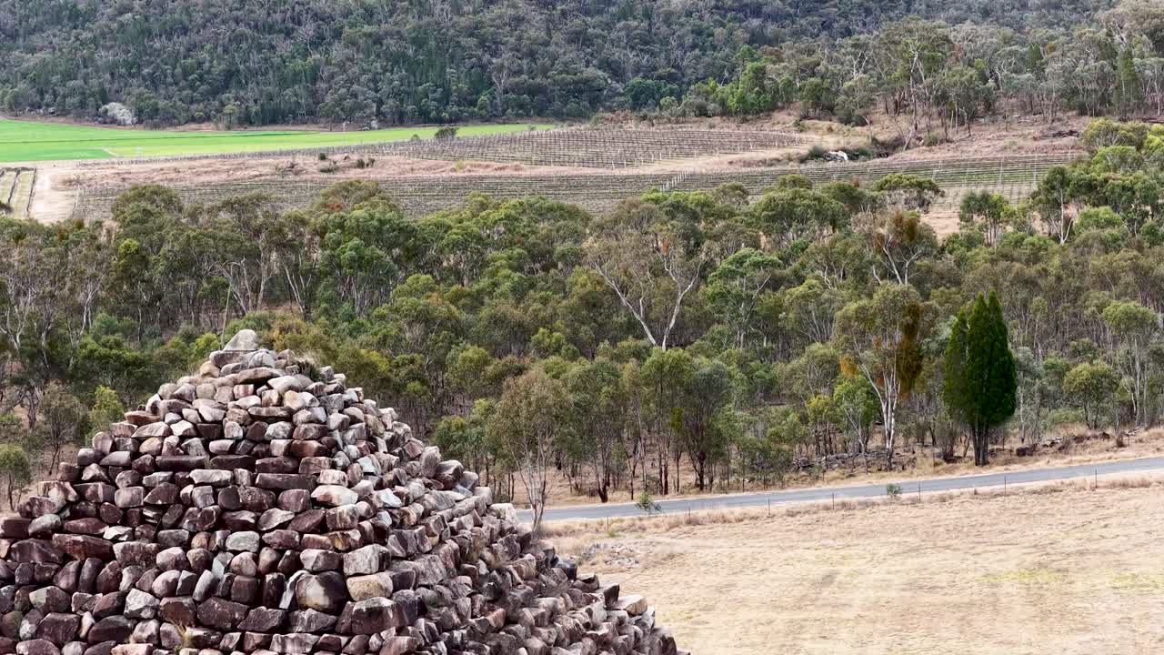 A stone pyramid stands in a dry, grassy field surrounded by bushland. The camera slowly pans right, revealing the rural Australian landscape under soft daylight