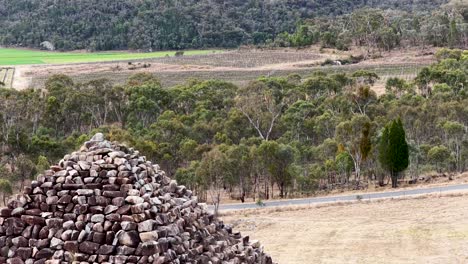 a stone pyramid stands in a dry, grassy field surrounded by bushland. the camera slowly pans right, revealing the rural australian landscape under soft daylight