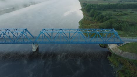 aerial establishing view of the steel bridge over lielupe river on a sunny summer morning, fog rising over the river, cars driving, wide drone dolly shot moving left