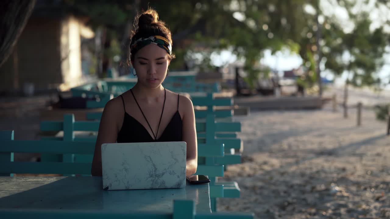 Young influencer girl working on laptop at beach lit by sunset, Bali