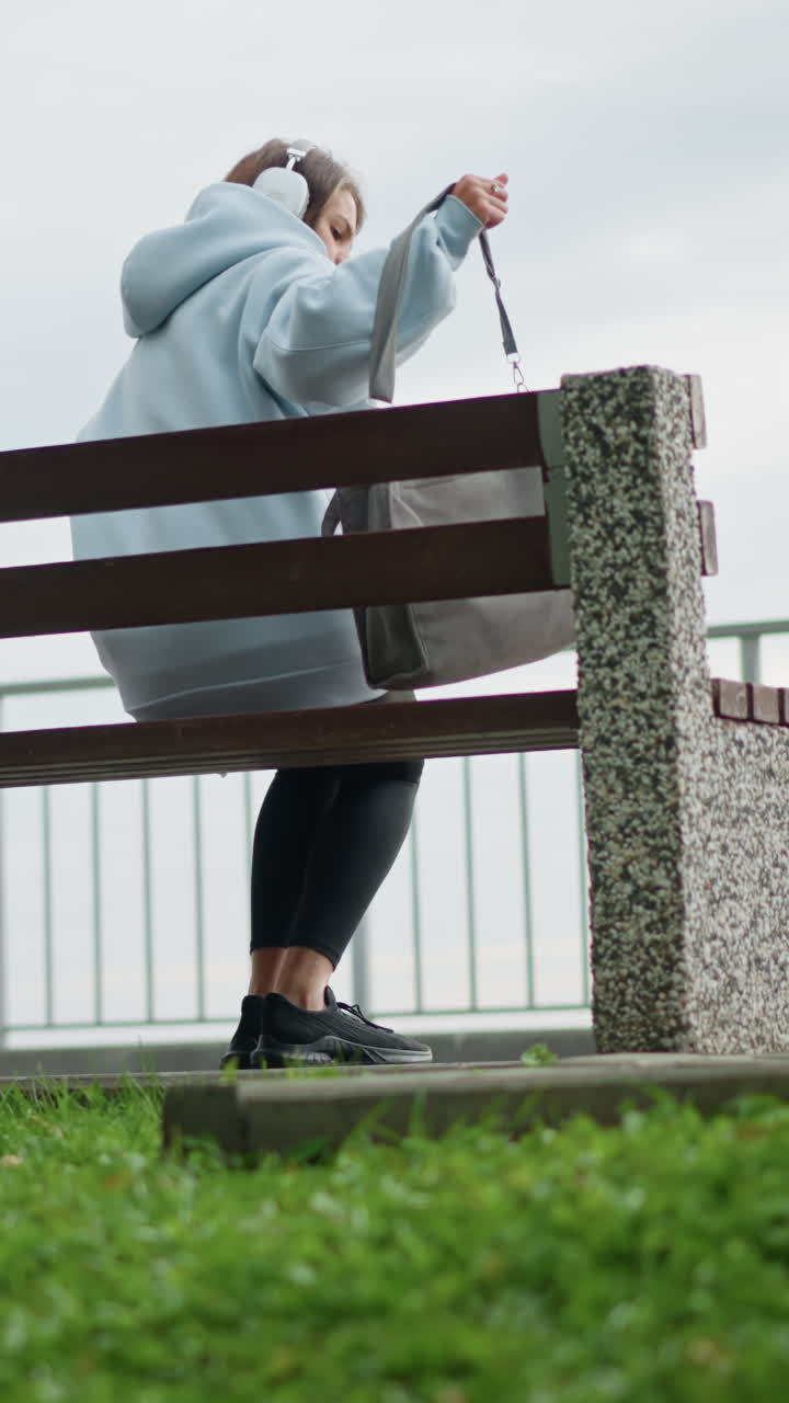 Pretty woman in casual wear walking gracefully toward concrete bench, sitting down and crossing her legs with bag beside her in serene outdoor environment, surrounded by grass and leaves