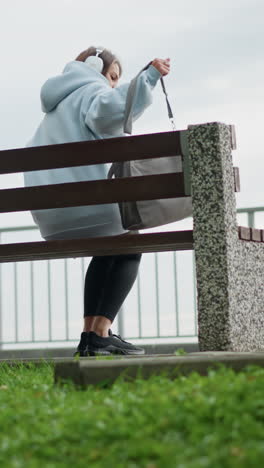 pretty woman in casual wear walking gracefully toward concrete bench, sitting down and crossing her legs with bag beside her in serene outdoor environment, surrounded by grass and leaves
