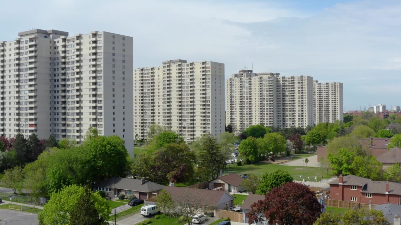 Riprese aeree ravvicinate con drone che mostrano molteplici torri residenziali che si ergono sopra un lussureggiante quartiere suburbano, ricco di alberi secolari e case unifamiliari, in una luminosa giornata di primavera.